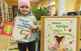 Uśmiechnięta dziewczynka w r&oacute;żowej czapce pokazuje sw&oacute;j dyplom i wypożyczoną książeczkę. Z prawej strony jest korkowa tablica z plakatem promującym projekt czytelniczy.