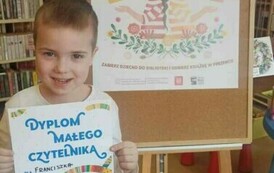 Uśmiechnięty chłopiec w blond włosach pokazuje sw&oacute;j otrzymany dyplom. Za nim stoi tablica korkowa z plakatem promującym akcję Mała Książka Wielki Człowiek