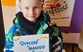 Chłopiec w blond włosach pokazuje sw&oacute;j otrzymany dyplom. Za nim stoi matalowa tablica z plakatem promującym projekt Mała Książka Wielki Człowiek.