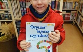 Chłopiec w blond włosach pokazuje sw&oacute;j otrzymany dyplom, uśmiecha się.