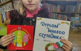 Dziewczynka w jasnej czapce stoi z dyplomem w ręku i wypożyczoną książeczeką.