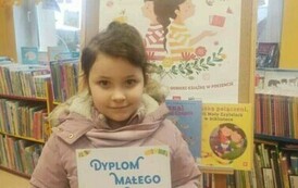 Dziewczynka w ciemnych włosach trzyma dyplom i kredki. Za nią jest tablica korkowa z plakatem promującym projekt Mała Książka Wielki Człowiek.