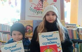 Dziewczynka i niższy chłopiec trzymają dyplomy. Dziewczynka ma jeszcze w ręku książeczkę, kt&oacute;rą wypożyczyła.Za nimi jest tablica korkowa z plakatem promującym projekt  Mała Książka Wielki Człowiek.