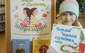 Dziewczynka trzyma dyplom, kartę Małego Czytelnika i wypożyczoną książeczkę. Z lewej strony stoi tablica korkowa z plakatem promującym projekt czytelniczy,