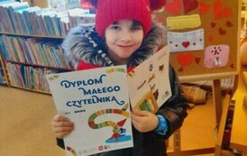 Dziewczynka w czerwonej czapce trzyma dyplom, kredki i tekturową kartkę z grą. Za nią tablica korkowa z walentynową grafiką.