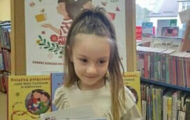 Uśmiechnięta dziewczynka o blond włosach trzyma książeczkę z wyprawki. Za nią stoi tablica korkowa z plakatem dotyczącym projektu Mała Książka Wielki Człowiek.