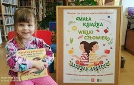 Dziewczynka w ciemnych wlosach siedzi trzymając nową książkę,a z prawej strony stoi korkowa tablica z plakatem dotyczącym projektu czytelniczego.