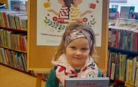 Dziewczynka w blond włosach w opasce i kamizelce trzyma książeczkę z wyprawki. Za nią jest korkowa tablica z plakatem projektu czytelniczego.