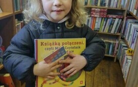 Dziewczynka w blond i lekko kręconych włosach trzyma w rękach książkę z wyprawki i kartę Małego Czytelnika.