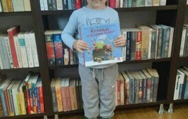 Chłopiec w ciemnych włosach pokazuje swoją książkę z wyprawki. Za nim regały z kisiążkami.