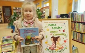 Dziewczynka o blond włosach trzyma książeczkę z wyprawki. Za nią stoi tablica korkowa z plakatem dotyczącym projektu Mała Książka Wielki Człowiek.