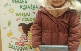 Uśmiechnięta dziewczynka o blond włosach trzyma książeczkę z wyprawki. Za nią stoi tablica korkowa z plakatem dotyczącym projektu Mała Książka Wielki Człowiek.