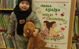Mały chłopiec stoi trzymająć w ręku brązowego misia. Za nim jest korkowa tablica z plakatem dotyczącym projektu czytelniczego.