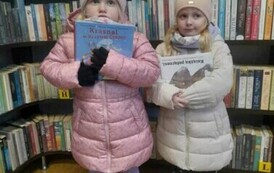 Dwie dziewczynki stoją z  książeczkami w rękach pokazując je. Za nimi są regały z książkami.