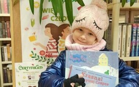 Dziewczynka stoi trzymając dwie książki, za nią jest korkowa tablica z plakatem dotyczącym projektu Mała Książka Wielki Człowiek