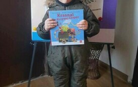 Chłopiec w kombinezonie i czapce stoi trzymając książkę z wyprawki. Za nim jest tablica metalowa z plakatem dotyczącym projektu czytelniczego.