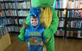 Duży zielony smok stoi z małym niebieskim smokiem, kt&oacute;ry trzyma książeczkę o zwierzątkach.