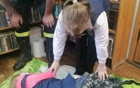 Dziewczynka leży na kocu bokiem udając poszkodowaną. Przy niej jest pochylona dziewczynka, kt&oacute;ra dotyka ją rękami,a z boku jest pochylony strażak.