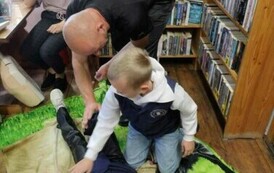 Dziewczynka leży na kocu udając poszkodowaną. Przy niej klęczy chłopiec patrząc na strażaka, kt&oacute;ry mu pokazuje co robić. Z tyłu widać na krześle nauczycielkę.