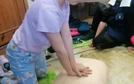 Dziewczynka w blond włosach uciska klatkę piersiową manekina. Obok widać chłopca, kt&oacute;ry na to patrzy.