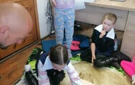 Dziewczynka pochylona jest nad manekinem dotykając jego bark&oacute;w, obok stoi inna dziewczynka. Z prawej strony siedzi chłopiec i patrzy, a z lewej strony siedzi strażak i obserwuje.