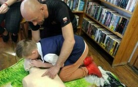 Chłopiec robi sztuczne oddychanie manekinowi, przy nim jest strażak, kt&oacute;ry palcem pokazuje mostek na menekinie.