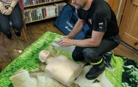 Na kocu jest połozony manekin, przy nim siedzi strażak i opowiada dzieciom o ratowaniu ludzi.