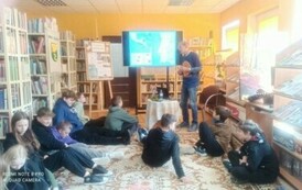 Młodziez siedzi na dywanie i słucha jak utor opowiada o swoich podr&oacute;żach. Na środku widać ekran dużego telewizora.