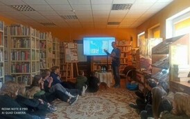 Młodzież siedzi na dywanie i patrzy na autora, kt&oacute;ry pokazuje coś na ekranie telewizora.