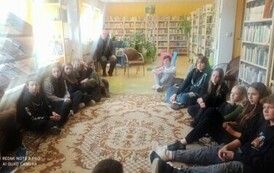 Młodzież siedzi na dywanie i patrzy w ekran telewizora, na kt&oacute;rym są wyświetlane zdjęcia z podr&oacute;ży autora.