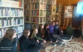 Młodzież siedzi na dywanie i patrzy w ekran telewizora, na kt&oacute;rym są wyświetlane zdjęcia z podr&oacute;ży autora.