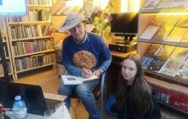 Autor podpisuje w książce dedykację dziewczynce, kt&oacute;ra kuca przy Nim. Przy autorze jest stolik z laptopem.