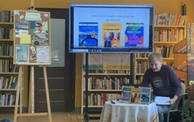 Przy okrągłym stoliku ze swoimi książkami siedzi podr&oacute;znik i opowiada, a na dużym ekranie są wyświetlane zdjęcia.