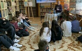 Młodzież siedzi na dywanie i słucha Michała podr&oacute;znika, kt&oacute;ry siedzi z przodu przy stoliku. Na dużym ekranie są wyświetlane zdjęcia.