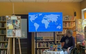 Przy okrągłym stoliku ze swoimi książkami siedzi podr&oacute;znik i opowiada, a na dużym ekranie są wyświetlane zdjęcia. Z lewej strony stoi tablica korkowa z grafikami o Tygodniu Bibliotek.