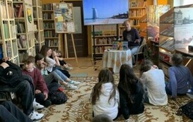 Młodzież siedzi na dywanie i słucha Michała podr&oacute;znika, kt&oacute;ry siedzi z przodu przy stoliku. Na dużym ekranie są wyświetlane zdjęcia.