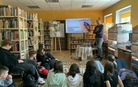 Młodzież siedzi na dywanie i słucha Michała podr&oacute;znika, kt&oacute;ry stoi z przodu przy stoliku. Na dużym ekranie są wyświetlane zdjęcia.