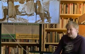 Przy okrągłym stoliku ze swoimi książkami siedzi podr&oacute;znik, a na dużym ekranie są wyświetlane zdjęcia. 