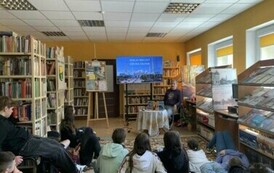 Młodzież siedzi na dywanie i słucha Michała podr&oacute;znika, kt&oacute;ry siedzi z przodu przy stoliku. Na dużym ekranie są wyświetlane zdjęcia.