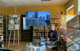 Przy okrągłym stoliku ze swoimi książkami siedzi podr&oacute;znik, a na dużym ekranie są wyświetlane zdjęcia. Z lewej strony stoi tablica korkowa z grafikami o Tygodniu Bibliotek.