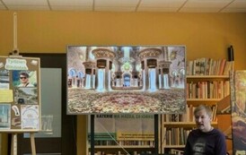 Przy okrągłym stolku ze swoimi książkami siedzi podr&oacute;znik, a na dużym ekranie są wyświetlane zdjęcia. Z lewej strony stoi tablica korkowa z grafikami o Tygodniu Bibliotek.