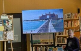 Przy okrągłym stolku ze swoimi książkami siedzi podr&oacute;znik, a na dużym ekranie są wyświetlane zdjęcia. Z lewej strony stoi tablica korkowa z grafikami o Tygodniu Bibliotek.