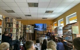 Młodzież siedzi na dywanie i słucha Michała podr&oacute;znika, kt&oacute;ry siedzi z przodu przy stoliku. Na dużym ekranie są wyświetlane zdjęcia.