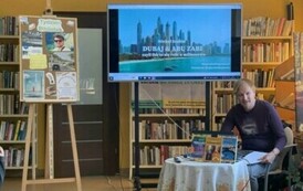 Przy okrągłym stolku ze swoimi książkami siedzi podr&oacute;znik, a na dużym ekranie są wyświetlane zdjęcia. Z lewej strony stoi tablica korkowa z grafikami o Tygodniu Bibliotek.