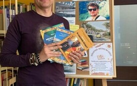 Michał Szulim stoi trzymając trzy swoje książki. Za Nim jest tablica korkowa z grafiką o Tygodniu Bibliotek.