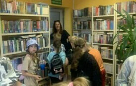 Dzieci stoją z placakami w bibliotece, w drzwiach stoi ich nauczycielka i się uśmiecha.