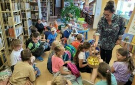 Dzieci siedzą w bibliotece na podłodze, bibliotekarka częstuje ich cukierkami z koszyczka.