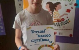 Dziewczynka w blond włosach pokazuje sw&oacute;j nowy dypolm. Za nią stoi metalowa tablica z plakatem dotyczącym akcji czytalniczej.