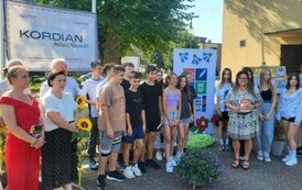 Przy banerze stoi grupa uczni&oacute;w z kierowniczką biblioteki i Radnymi, kt&oacute;rzy trzymają słoneczniki.