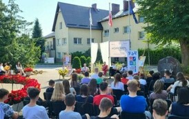 Widok z daleka na siedzącą publiczność i czytających Radnych. Po środku jest baner, po lewej stronie firanki,a po prawej rolka z logiem biblioteki.